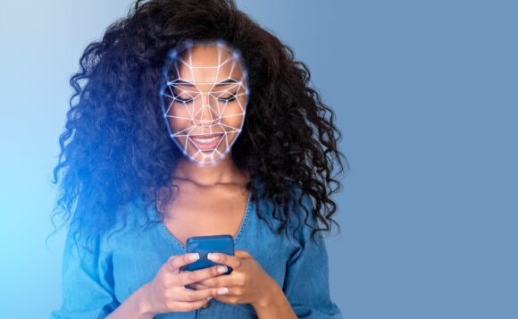A Black woman in a blue shirt smiling while she types on her phone, her face covered in a web of lines mapping it out